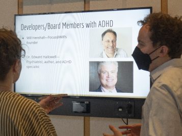 Matthew Osterholzer stands in front of a large monitor displaying his project, explaining information to an audience member.